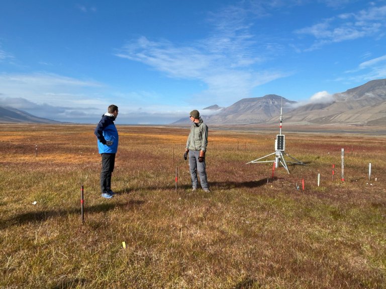Ministeren og forskeren ser utover tundraen. Fjell i bakgrunnen. Målestasjonen til høyre.