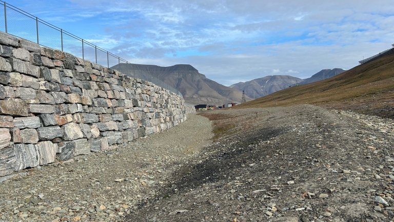 En høy skredvoll bygget i store steiner, stålgjerde på toppen. Fjell i bakgrunnen. 