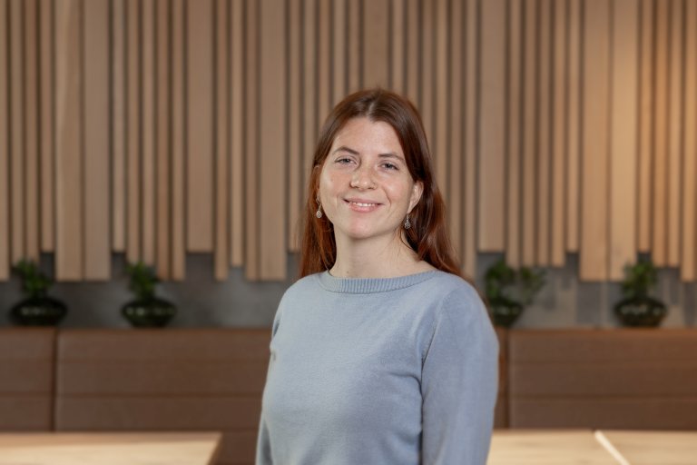 Halvportrett av kvinne, brunt, langt hår og grå genser. Står foran en spilevegg.
