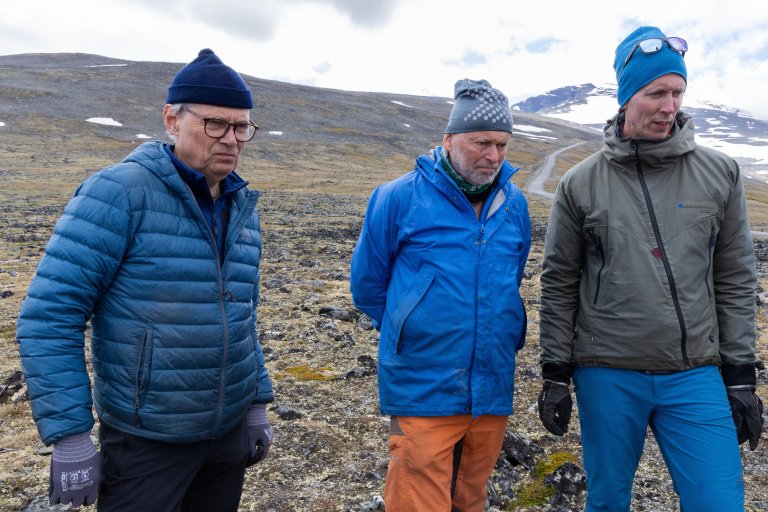Tre menn står midt i fjellheimen og ser litt ned og til høyre