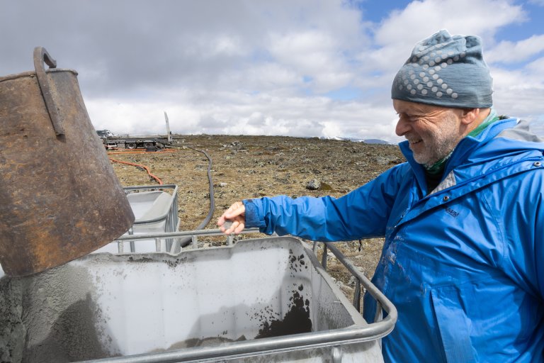 Bernd står med blå lue og jakke og holder sand i hånda over en hvit, stor boks.