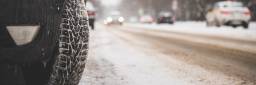 Bildet har fokus rettet mot på et piggdekk til en bil som står parkert langs en trafikkert vei. Veien er delvis dekket av snø.
