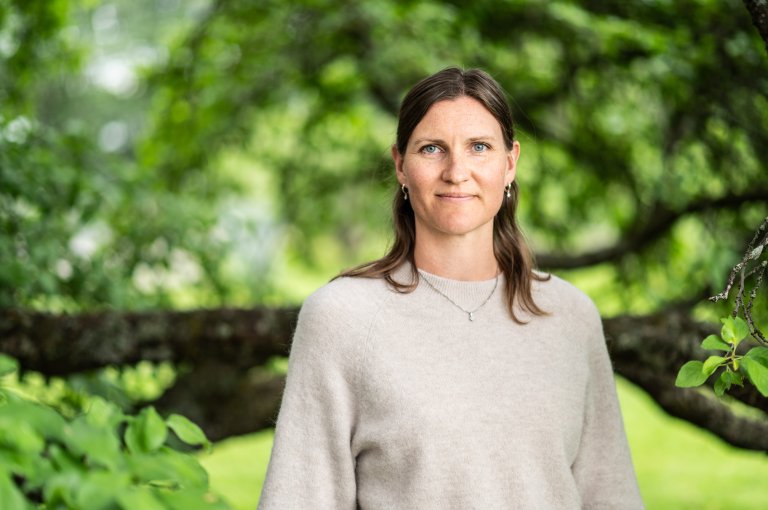 Halvportrett kvinne ute, foran et stort og grønt tre.