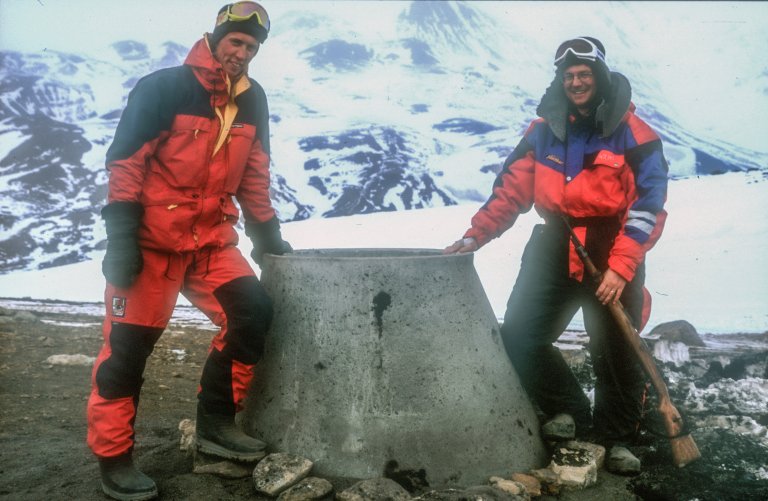 Vinterkledd Ketil Isaksen og Daniel Vonder Mühll står på hver sin side, av en betongkloss som er toppen av permafrost-stasjonen. Bak skimtes fjell delvis dekket av snø.
