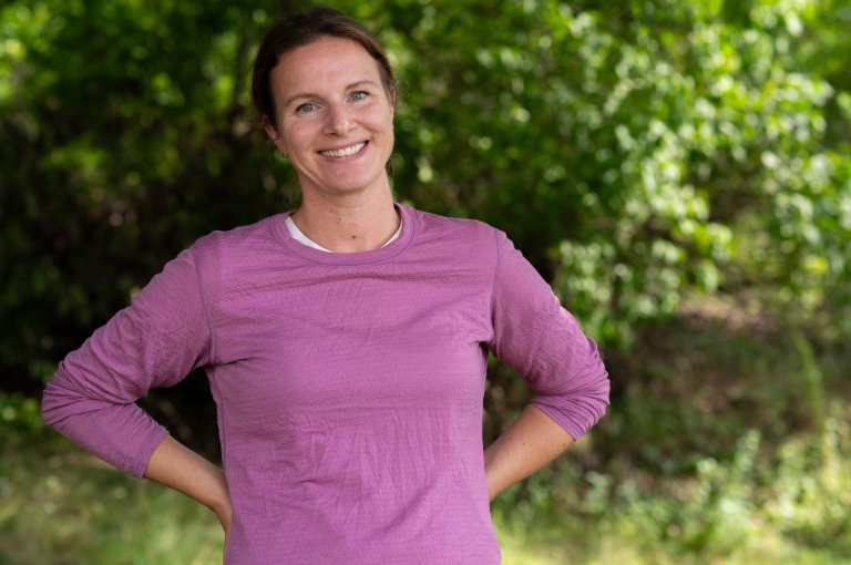Halvportrett kvinne, rosa ulltrøye, ute og foran en grønn "vegg" av vegetasjon.
