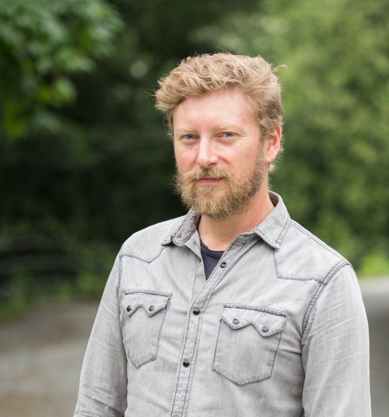 Øyvind med skjegg og grå olaskjorte står foran duse, grønne trær-