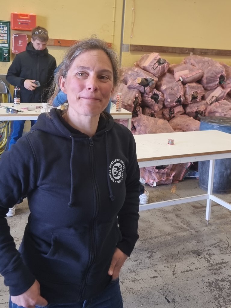 Lena, med en hettegenser med logo "Arven etter Nansen" på, smiler foran en ung gutt i bakgrunnen, i et lokale med vedstabler og skrivebord.