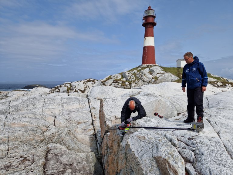 Et fyrly i bakgrunnen av to fyrer fra MET som jobber med verktøy, en sitter på kne og han andre står oppreist og følger med.