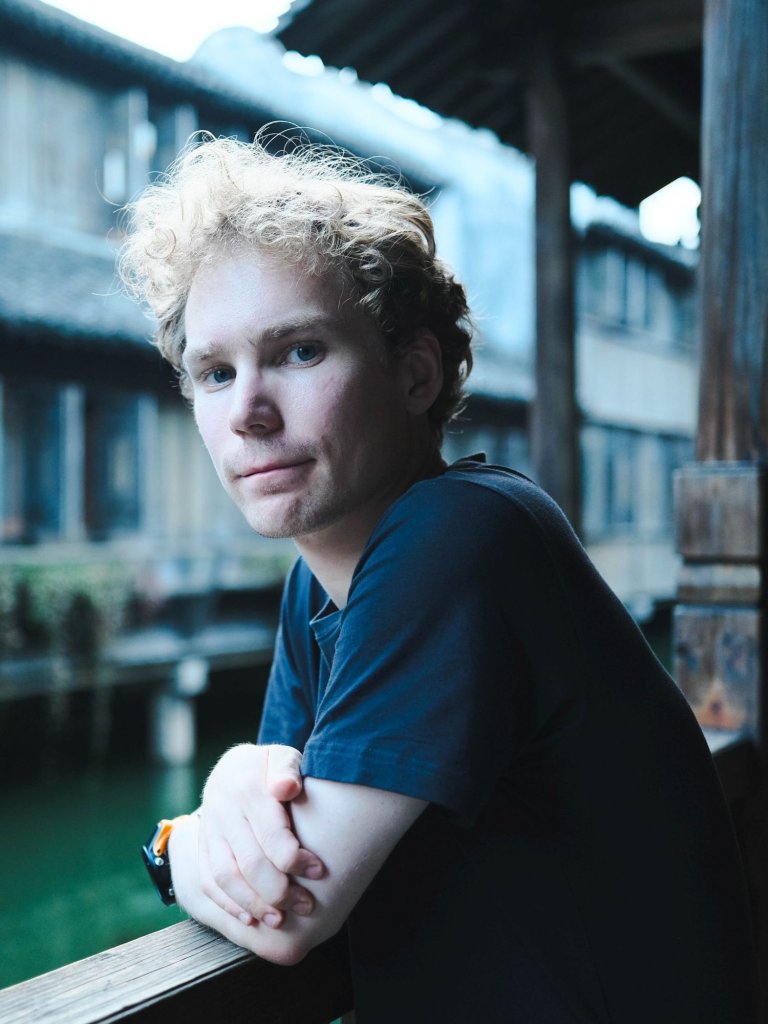 Halvportrett mann med t-skjorte ute, lener seg på et gjerde med armer i kryss.